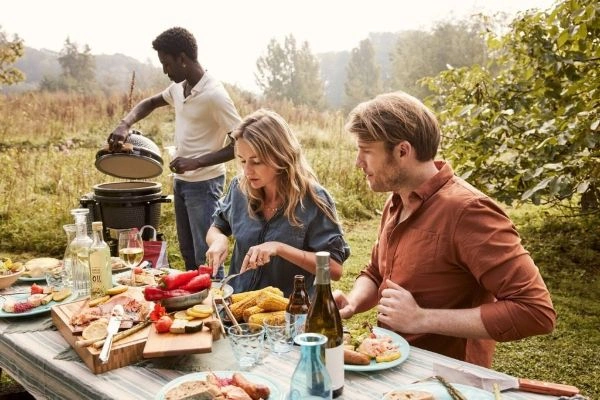 Doua persoane ce mananca la masa si o persoana langa un gratar Grill Guru Original XL, afara in natura.